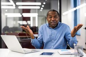 Businessman in blue shirt sitting at desk with laptop and tablet, gesturing confusion in modern office. Concept of frustration, problem-solving, misunderstanding, and communication issues workplace. photo
