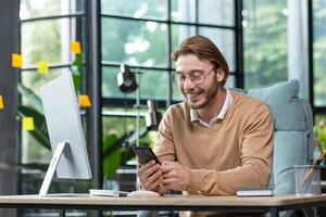 A smiling young businessman sits in the office at a desk and uses a mobile phone, writes a message, searches for information on the Internet, checks social networks. photo