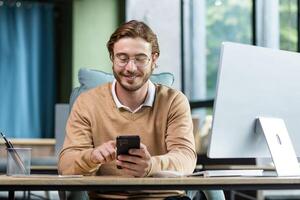 A young freelancer, programmer works from home. Sits at the desk and uses the phone, writes messages, checks mail. photo