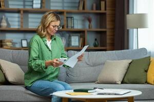 Senior beautiful woman in glasses sitting on sofa at home. Worriedly holding the phone and documents in his hands. Problem with accounts, credit, payment. photo