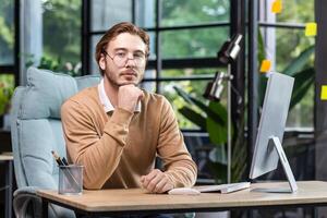 Portrait of thinking serious man with computer inside modern green loft office, blond man looking thoughtfully at camera businessman in casual clothes working with computer. photo