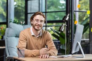 Portrait of successful smiling programmer inside modern green loft office, blond man smiling and looking at camera, businessman in sweater and casual shirt close up working with computer. photo