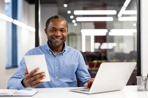 Smiling businessman using tablet and laptop in modern office setting. Confident professional enjoying productive work environment. Happy expression, business concept, successful professional life. photo