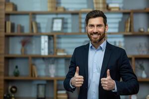 Caucasian businessman in suit giving thumbs up gesture while standing on background of office. Successful male making positive and approving sign with both hands while looking at camera indoors. photo