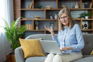 Senior woman sitting on a sofa, frustrated with computer problems while working from home. She looks confused and is gesturing with her hand, indicating technical issues with her laptop. photo