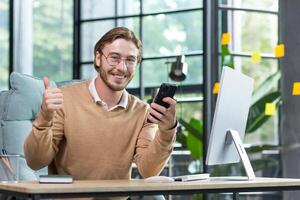 A young freelancer, programmer, student works from home. Sits at the desk and uses the phone, writes messages, checks mail. He looks at the camera, points a super finger. photo