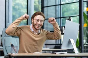 A young businessman, student, freelancer, programmer sits in the office at the table and celebrates success, victory. Joyfully raises his hands up and shows strength, looks into the camera, smiles. photo