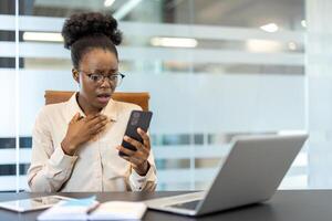 Businesswoman looking shocked while checking smartphone in modern office. Surprised expression on woman's face while sitting at desk with open laptop and documents. photo