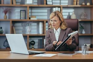 Online business meeting. A senior woman in a suit is sitting in the office at a desk and working on a laptop, talking on a call and anxiously showing documents to the camera. photo