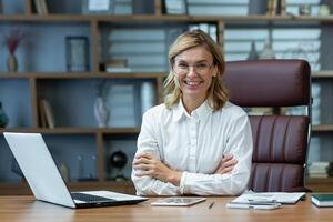 Successful businesswoman boss in shirt sitting at table with crossed arms smiling and looking at camera, lawyer working in modern office using laptop at work. photo