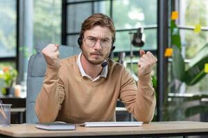 An angry young man, a businessman, a programmer in a headset, sits in the office and speaks expressively to the camera, holds his fists in his hands. Online meeting, discussion, deadline, project. photo
