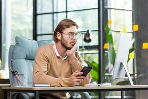 Upset man sitting at the table with a phone in his hands looks tiredly at the computer screen, holds his head with his hand, overworked burnout. photo