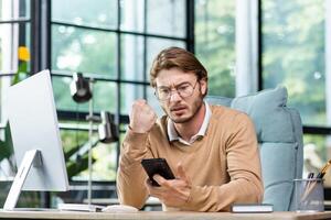 A young man businessman sits in the office at the table and solves questions, problems on the phone. Worriedly looks at the phone, worries, reads messages, checks bills. photo