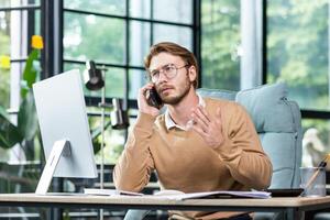 A young man businessman sits in the office at the table and solves questions, problems on the phone. Talks, gesticulates worriedly with his hands, explains. photo