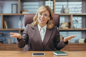 Serious and worried senior business woman in suit sitting at desk in office and talking to camera on call, gesturing and throwing hands in frustration. photo