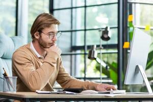 A young businessman sits in the office at a table, works with documents, looks at notes in a notebook, thinks about a strategy, analyzes. He rubs his beard thoughtfully with his hand. photo