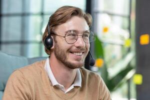 Successful businessman with call headset working inside office, man consulting online clients, talking with colleagues on online remote meeting, sitting at workplace with laptop photo
