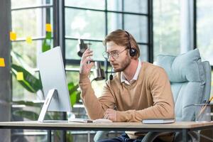 Business online meeting. A serious young man, a businessman, a programmer sits in the office in a headset and talks on a call from a computer. Communicates with clients, partners. Gesturing with hands. photo