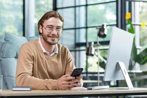 Friendly businessman in glasses is working in the office at the table, holding the phone in his hands, texting, smiling and looking at the camera. photo