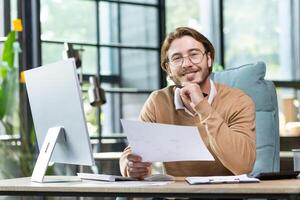 Successful and confident young man businessman, freelancer works in a modern office with documents, papers. Sitting at a desk with a computer. Smiling looking at the camera. photo