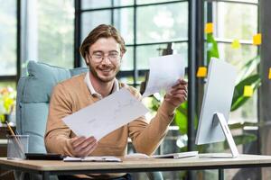 A young businessman, a freelancer works in a modern office with documents and papers. Sitting at a desk with a computer. photo