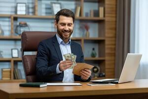 Photo of a happy young businessman sitting at a desk in the office and holding an envelope with cash in his hands, looking satisfied at the received banknotes.