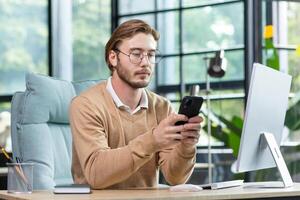 A young freelancer, programmer works from home. Sits at the desk and uses the phone, writes messages, checks mail. photo