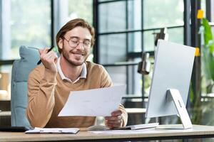 Satisfied young guy doing paperwork in the office, sitting at a desk in front of a computer, joyfully going through business papers, smiling photo