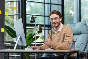 A young male businessman is concentrating on working from a home office at a computer. Smiles at the camera, points a super finger. photo
