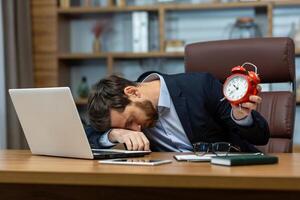 Tired office manager laying down on folded hand on table with personal gadgets and holding red alarm clock. Overworked man having insomnia or chronic fatigue syndrome and taking nap during busy day. photo
