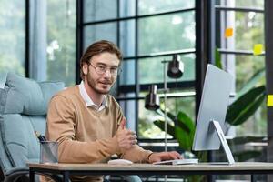 Portrait of a young man in glasses working at a computer in a modern office. He looks at the camera, smiles, shows a successful gesture with a super finger. photo