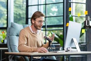 A young male teacher who sits in the office at a table with a computer and conducts online lessons, lectures, training. Speaks into the microphone, tells, explains. photo