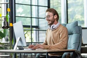 Young smiling male student in headphones sits on campus at a desk and studies online at a computer. Writes coursework and diploma work. photo
