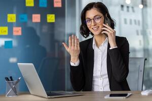 Confident businesswoman talking on phone while using laptop in a modern office. Professional attire and positive communication. photo
