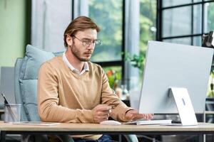 Concentrated young man holds a card in his hands, sits at a desk in the office in front of a computer, makes an online payment, purchases online. photo