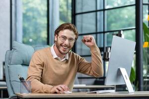 A happy young male student sits in the office, at home in front of the computer, shows a winning gesture of success with his hand, celebrates, looks at the camera smiling. photo