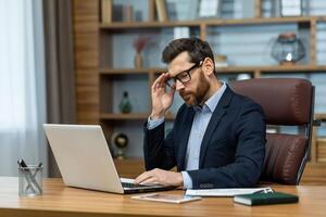 Frustrated administrator experiencing headache while sitting by desk with portable computer in modern interior. Exhausted man feeling pain in temple and getting distracted from working process. photo