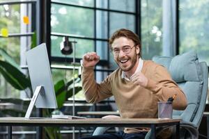 Very happy businessman raises his hands up, rejoices at good news, sits at the desk in the office in front of the computer, looks at the camera, smiles. photo