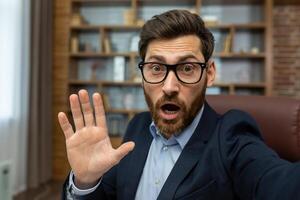 Mature man in a business suit appears shocked or overwhelmed, gesturing with his hand as if saying no or stop while in a messy home office setting. photo