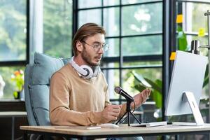 A young male blogger is sitting in a home office at a desk with a computer in front of a microphone and wearing headphones. Records podcast, blog, communicates online via call, webinar. photo