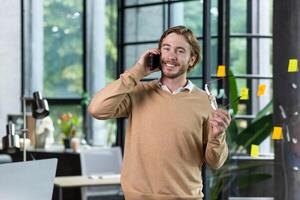 A young freelancer man is standing in the office and talking on the phone. He holds glasses in his hands, looks at the camera smiling. photo