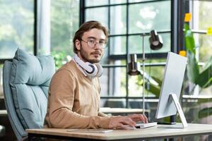 Portrait of a serious office worker with headphones around his neck sitting at a desk, working at a computer, tapping on a keyboard, looking seriously at the camera. photo