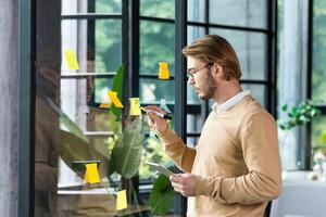 A young man in casual clothes stands near a transparent note board and attaches stickers with reminders and plans to it, the concept of work planning in the office. photo