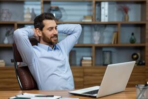 Relaxed brunette man leaning back and holding hands behind head in personal workspace with pc. Calm head of company taking break during busy day and enjoying daydreaming in comfortable chair. photo
