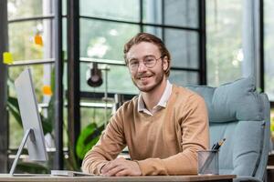 Portrait of a successful young man, programmer, IT developer, software sitting at a desk in a modern office, working at a computer. He looks at the camera, smiles. photo