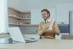 Young teacher teaches remotely from home, man works in kitchen at home, uses laptop for online call with communicates with students smiling explains assignment shows notebook. photo