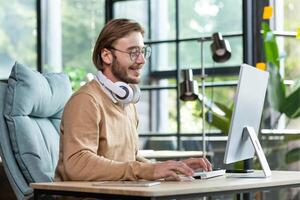 Young smiling blond male student in headphones, beige sweater and glasses. Sitting at the table, studying at the computer. Writes on the keyboard, talks on a call, listens to lectures. photo