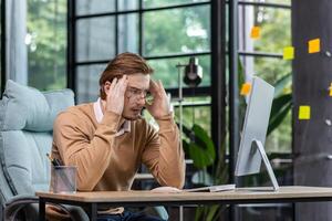 A young male programmer, IT developer codes, thinks about a new project. He is sitting concentrated at the desk in the office, holding his head thoughtfully with his hands, looking at the monitor. photo