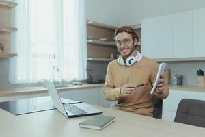 Young teacher teaches remotely from home, man works in kitchen at home, uses laptop for online call with communicates with students smiling explains assignment shows notebook. photo