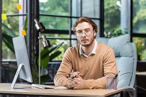 Portrait of thinking serious man with computer inside modern green loft office, blond man looking thoughtfully at camera businessman in casual clothes working with computer. photo